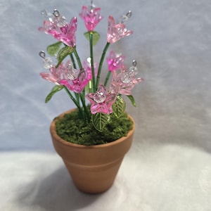 May include: A small terracotta pot filled with green moss, holding a bouquet of pink and clear glass flowers. The flowers have green stems and leaves, with a star-shaped flower in the center. The flowers are a mix of light and dark pink.