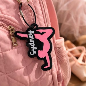 May include: A pink backpack with a gold zipper and a black and pink keychain. The keychain features a ballerina silhouette with the name "Sydney" in white script. The background includes ballet shoes.