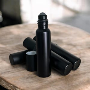 May include: A collection of black roller bottles with a matte finish. One bottle stands upright, with a gray rollerball top. Several other bottles and caps are scattered around on a wooden surface. The bottles are cylindrical and appear to be empty.