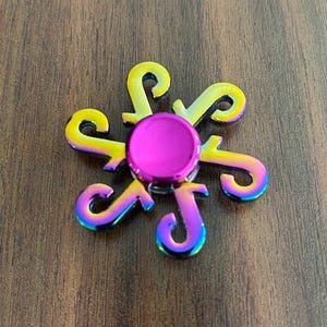 May include: A colorful fidget spinner with a flower-like design. The center is a bright pink circle, and the six curved arms transition from yellow to blue with a rainbow effect. The spinner is on a brown wooden surface.