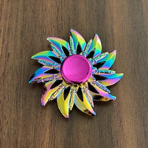May include: A colorful flower-shaped fidget spinner with a vibrant, iridescent finish. The spinner features a bright pink center button and petal-like blades in shades of blue, green, and yellow. The toy is designed for stress relief and entertainment.