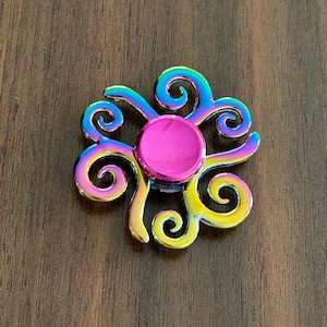 May include: A colorful fidget spinner with a flower-like design. The center is a bright pink circle, surrounded by swirling, iridescent arms in shades of blue, yellow, and purple. The spinner sits on a dark wood surface.