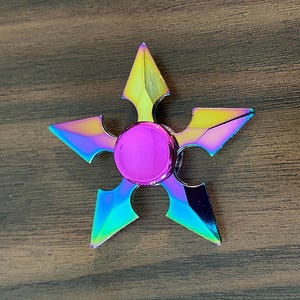 May include: A five-pointed fidget spinner with a rainbow iridescent finish. The center is a bright pink circle. The points are shaped like arrowheads. The spinner is on a brown wooden surface.