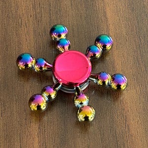 May include: A metallic fidget spinner with a vibrant rainbow finish. The spinner features a central pink hub and six arms, each ending in a cluster of three iridescent spheres. The toy is designed for stress relief and entertainment.