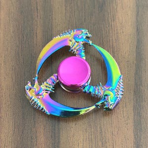 May include: A multi-colored fidget spinner with a central pink button and three curved blades. The blades have a rainbow iridescent finish with a dragon-like design. The spinner is designed to be held and spun by hand.