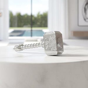 May include: A silver-colored hammer with a detailed design, resting on a white marble surface. The hammer's head features engraved text and a circular emblem. The handle has a textured, ribbed design. The hammer is a replica of Thor's hammer.