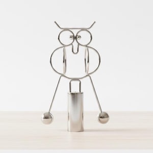 May include: A silver-colored owl-shaped napkin holder. The owl is constructed from bent metal rods and features large circular eyes, a beak, and a cylindrical body. Two spherical balls serve as feet. The napkin holder is designed to stand upright on a flat surface.