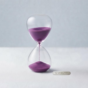 May include: A clear glass hourglass filled with vibrant purple sand, showing a portion of the sand flowing through the center. A small, rectangular silver-toned metal tag is placed next to the hourglass, clearly engraved with the text 15 MINUTES.