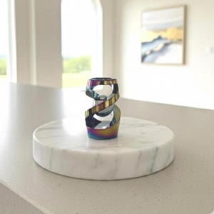 May include: A decorative, iridescent metal sculpture in a spiral design. The sculpture sits on a round, white marble base. The piece has a rainbow finish, reflecting various colors. The sculpture is a modern home decor item.