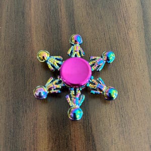 May include: A multi-colored fidget spinner with a central pink circle and six astronaut-shaped arms. The arms have a rainbow metallic finish, and the center is a bright pink color. The spinner is designed to be held and spun.