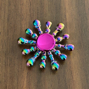 May include: A metallic fidget spinner with a vibrant, iridescent finish. The spinner features a central pink hub and radiating arms with rounded ends, displaying a spectrum of colors like blue, green, and purple. The design is symmetrical and sits on a wooden surface.