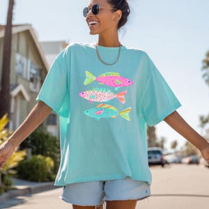 May include: A light blue t-shirt featuring three colorful fish illustrations. The fish have pink, blue, and patterned designs with yellow and orange fins. The shirt is a relaxed fit, suitable for casual wear.