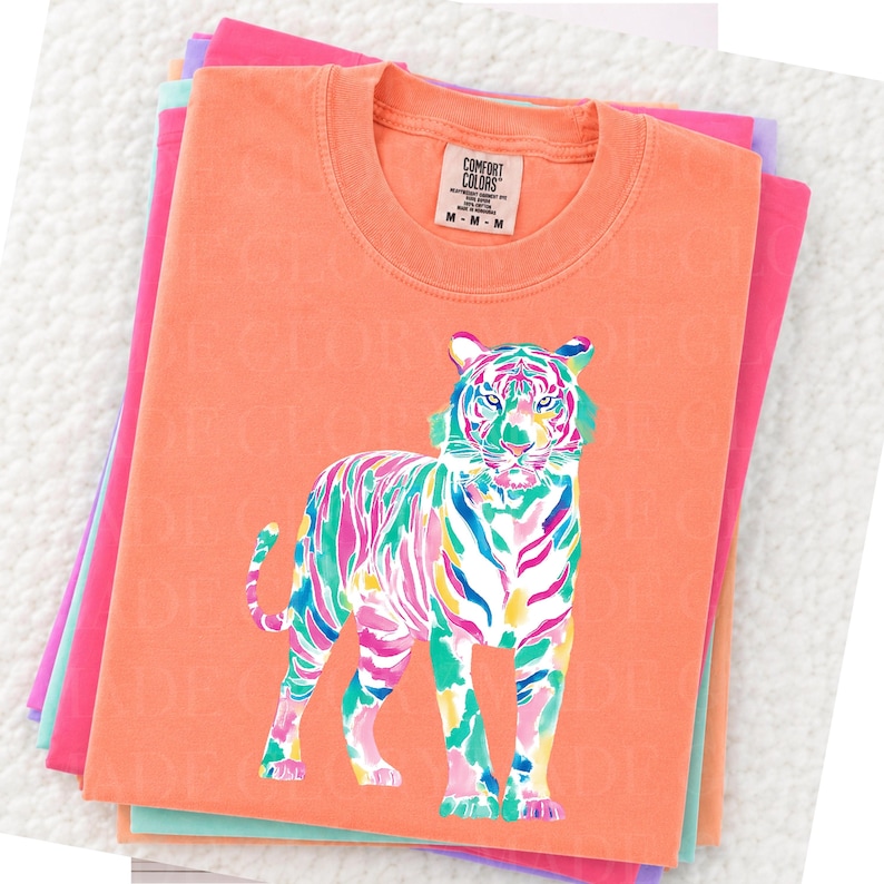 May include: Coral t-shirt with a colourful tiger illustration. The tiger design uses vibrant pink, blue, green, and yellow hues. The shirt has a crew neck and short sleeves. The tag reads "Comfort Colors" and "M-M-M".
