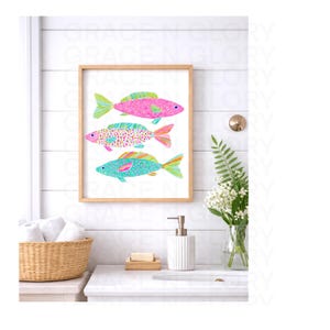 May include: A framed print featuring three colorful fish illustrations. The fish are pink, turquoise, and purple with patterned details and bright fins. The artwork is displayed in a bathroom setting with a white sink, towels, and decorative items.