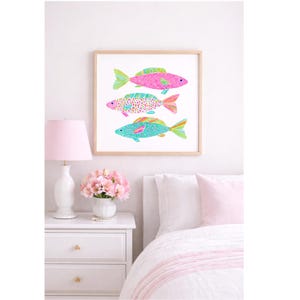 May include: A framed art print featuring three colorful fish illustrations. The fish are pink, turquoise, and patterned with dots and stripes. The artwork is displayed above a white nightstand with a pink lamp and flowers, creating a bright and cheerful bedroom scene.