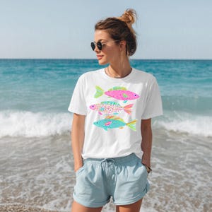 May include: White t-shirt featuring three colorful fish designs in pink, purple, and turquoise. The fish have patterned scales and fins. The person is wearing sunglasses and blue shorts, standing on a beach with the ocean in the background.