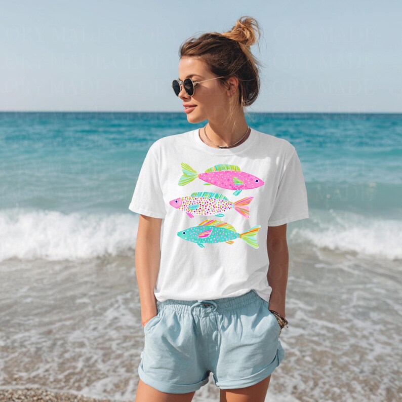 May include: White t-shirt featuring three colorful fish designs in pink, purple, and turquoise. The fish have patterned scales and fins. The person is wearing sunglasses and blue shorts, standing on a beach with the ocean in the background.