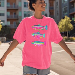 May include: Bright pink t-shirt featuring three colorful fish illustrations. Each fish has a unique pattern of pink, blue, yellow, and green, with a detailed design. The shirt is a comfortable, oversized fit, perfect for casual wear.