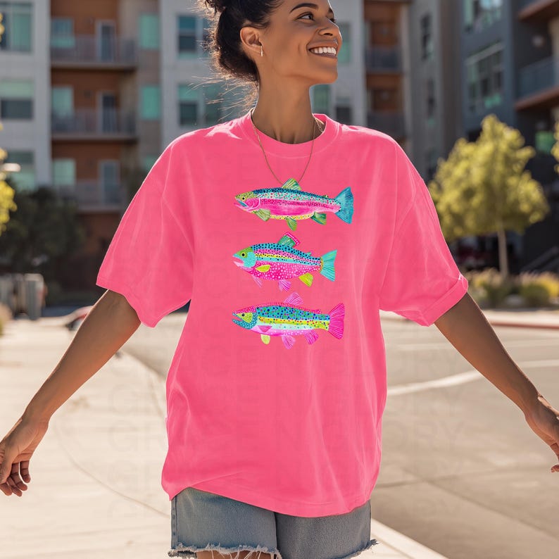 May include: Bright pink t-shirt featuring three colorful fish illustrations. Each fish has a unique pattern of pink, blue, yellow, and green, with a detailed design. The shirt is a comfortable, oversized fit, perfect for casual wear.