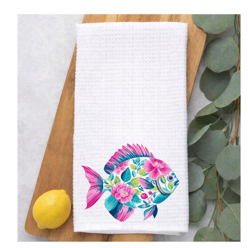 May include: White waffle-weave kitchen towel featuring a colorful fish design. The fish is adorned with pink, blue, and green floral patterns. A lemon and greenery are in the background.