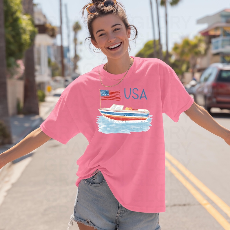 Op de afbeelding: Een roze t-shirt met een afbeelding van een boot, een Amerikaanse vlag en het woord "USA" in blauw. De boot heeft rood-wit-blauwe strepen. Het shirt heeft een losse pasvorm.