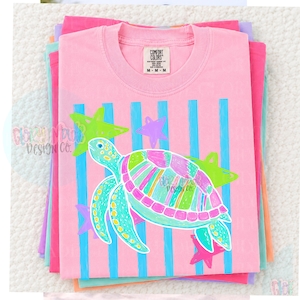 May include: A light pink short-sleeve t-shirt featuring a graphic of a sea turtle with a multi-colored shell in shades of pink, green, and blue. The design includes vertical blue stripes and several star shapes in lime green and pink.