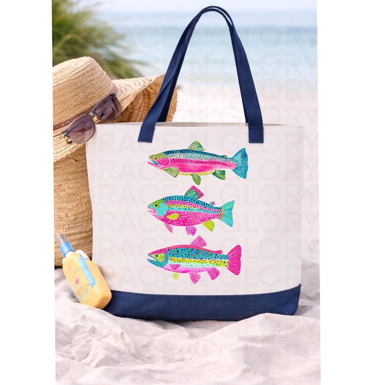 May include: White canvas tote bag with a navy blue base and handles. The bag features three colorful fish illustrations in shades of pink, blue, and green. A straw hat, sunglasses, and sunscreen are in the background.