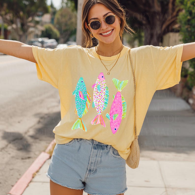 May include: Yellow t-shirt featuring three colorful fish illustrations. The fish are in shades of blue, pink, and purple with patterned details. The shirt is a relaxed fit and is paired with denim shorts.