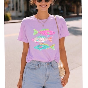 May include: A light purple t-shirt featuring three colorful fish designs. The fish have pink, blue, and green accents, with patterned scales. The shirt is short-sleeved and has a relaxed fit, suitable for casual wear.