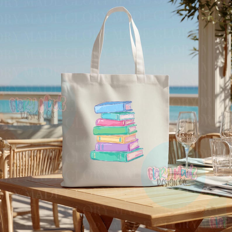 May include: A cream-colored tote bag with a stack of colorful books. The books are in shades of blue, pink, green, and purple. The bag has white handles and the text "GLORY MADE DESIGN CO." is visible.