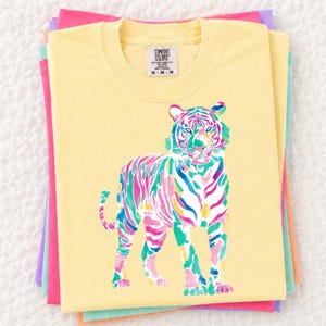 May include: A stack of pastel-coloured t-shirts. The top shirt is yellow with a colourful tiger graphic. The tiger is depicted in shades of pink, blue, green, and yellow. The shirt has a crew neck and the brand name "Comfort Colors" is visible.