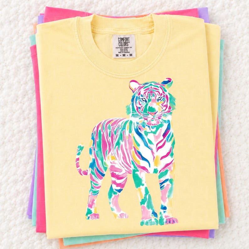 May include: A stack of pastel-coloured t-shirts. The top shirt is yellow with a colourful tiger graphic. The tiger is depicted in shades of pink, blue, green, and yellow. The shirt has a crew neck and the brand name "Comfort Colors" is visible.