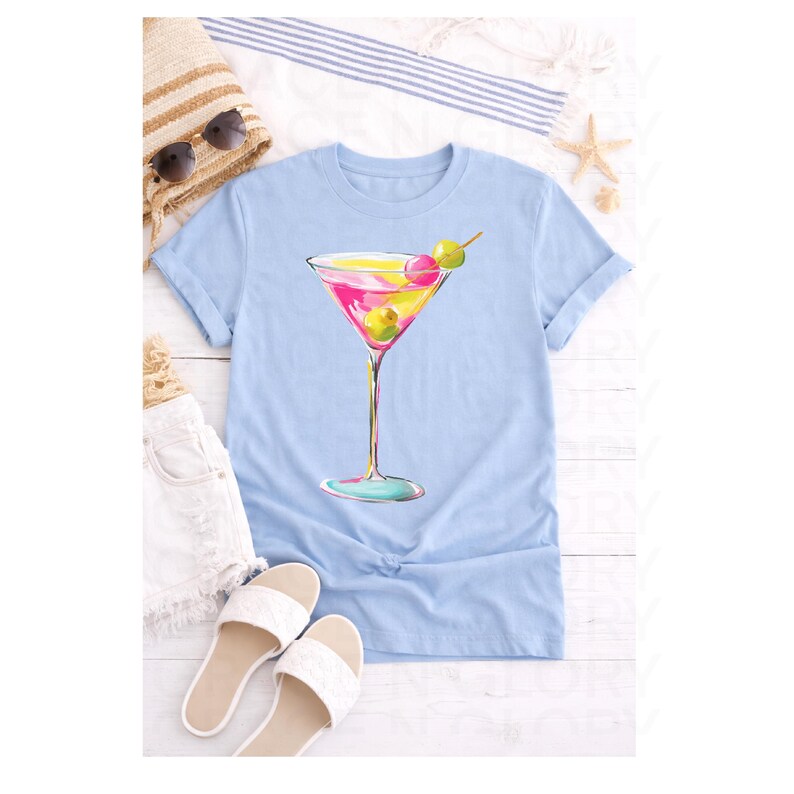 May include: A light blue t-shirt featuring a colorful cocktail illustration. The artwork depicts a martini with pink, yellow, and green accents, complete with olives. The shirt is styled with white shorts, sandals, and a straw tote bag.