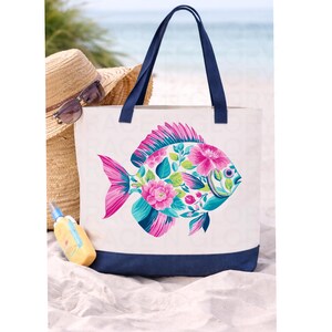 May include: White canvas tote bag with a navy blue base and handles. The bag features a colorful fish design filled with pink and teal flowers and leaves. A straw hat, sunglasses, and sunscreen are in the background.