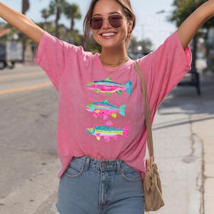 May include: A pink t-shirt featuring three colorful fish designs. Each fish has a unique pattern with shades of blue, pink, yellow, and green. The shirt is made of a soft material and has a relaxed fit.