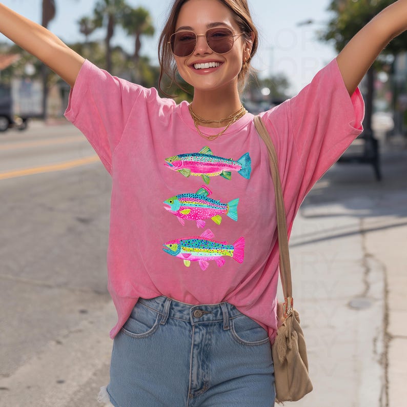 May include: A pink t-shirt featuring three colorful fish designs. Each fish has a unique pattern with shades of blue, pink, yellow, and green. The shirt is made of a soft material and has a relaxed fit.