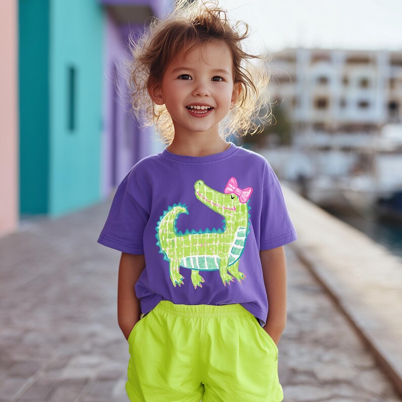 May include: A purple t-shirt featuring a green alligator with a pink bow. The alligator design has a checkered pattern. The child is wearing neon green shorts. The background has colorful buildings.