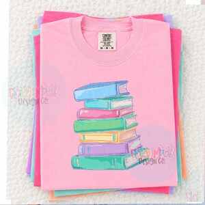 May include: A pink Comfort Colors t-shirt with a stack of colorful books graphic. The books are in shades of blue, pink, green, yellow, and purple. The shirt is folded and stacked with other pastel-colored shirts.