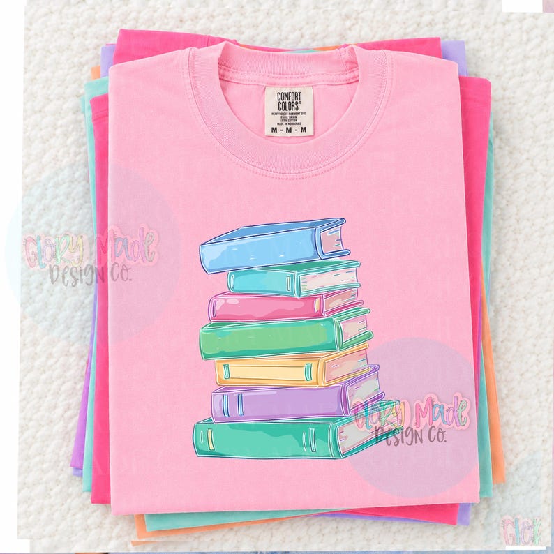 May include: A pink Comfort Colors t-shirt with a stack of colorful books graphic. The books are in shades of blue, pink, green, yellow, and purple. The shirt is folded and stacked with other pastel-colored shirts.