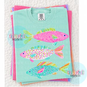 May include: A light teal Comfort Colors t-shirt featuring three colorful fish illustrations. The fish are pink, teal, and orange with dotted and striped patterns. The shirt is stacked on top of other folded shirts in pink, purple, and orange.