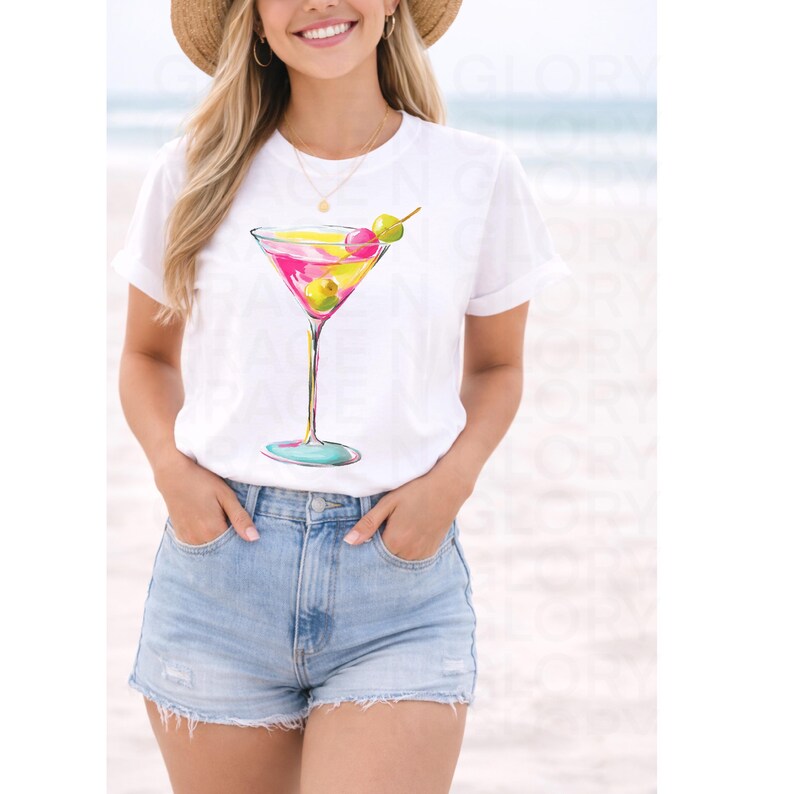 May include: White t-shirt featuring a watercolor illustration of a martini cocktail. The cocktail glass has pink and yellow liquid, green olives, and a colorful stem. The shirt is paired with denim shorts.
