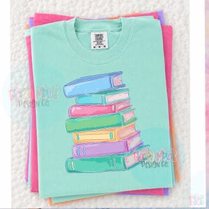 May include: A stack of colorful books is printed on a light teal Comfort Colors t-shirt. The books are in shades of blue, pink, green, yellow, and purple. The shirt is folded on top of other folded shirts in pink, purple, and orange.