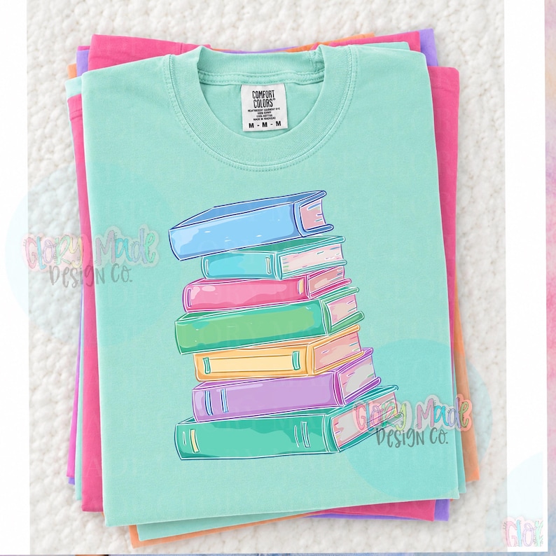 May include: A stack of colorful books is printed on a light teal Comfort Colors t-shirt. The books are in shades of blue, pink, green, yellow, and purple. The shirt is folded on top of other folded shirts in pink, purple, and orange.