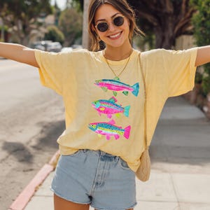 May include: Yellow t-shirt featuring three colorful fish illustrations. The fish have rainbow-colored bodies with blue, pink, and green accents. The shirt is paired with denim shorts. The overall style is casual and summery.