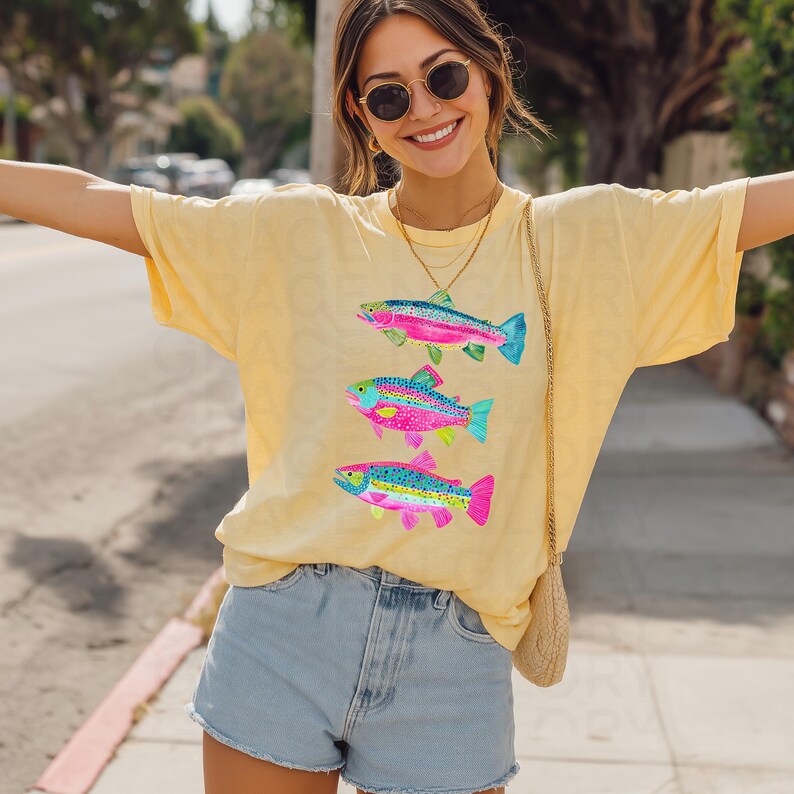 May include: Yellow t-shirt featuring three colorful fish illustrations. The fish have rainbow-colored bodies with blue, pink, and green accents. The shirt is paired with denim shorts. The overall style is casual and summery.