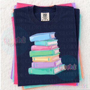 May include: Navy blue t-shirt with a stack of colorful books graphic. The books are in shades of blue, pink, green, yellow, and purple. The shirt is folded on top of other colorful shirts. The tag reads "Comfort Colors".