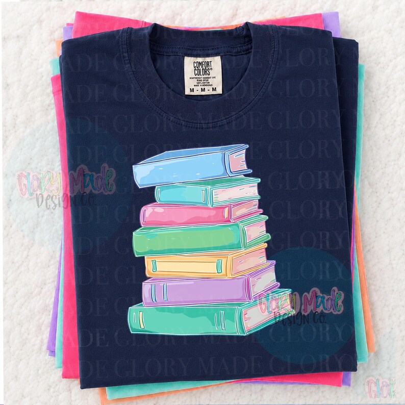 May include: Navy blue t-shirt with a stack of colorful books graphic. The books are in shades of blue, pink, green, yellow, and purple. The shirt is folded on top of other colorful shirts. The tag reads "Comfort Colors".