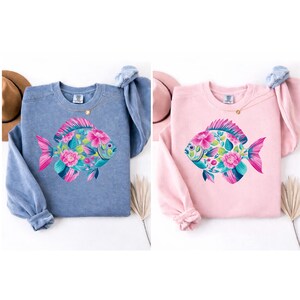 May include: Two sweatshirts, one blue and one pink, each featuring a colorful fish design. The fish is adorned with pink flowers and green leaves. The sweatshirts have long sleeves and a crew neck.