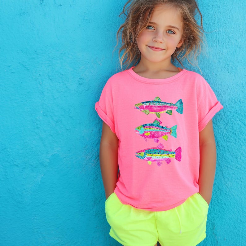 May include: A young person wearing a bright pink t-shirt with three colorful fish designs. The fish have blue, green, pink, and yellow accents. The person is also wearing neon yellow shorts, standing in front of a blue wall.
