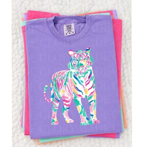 May include: A lilac Comfort Colors t-shirt with a colourful tiger illustration. The tiger is rendered in bright pink, green, blue, and yellow. The shirt is stacked with other brightly coloured shirts.
